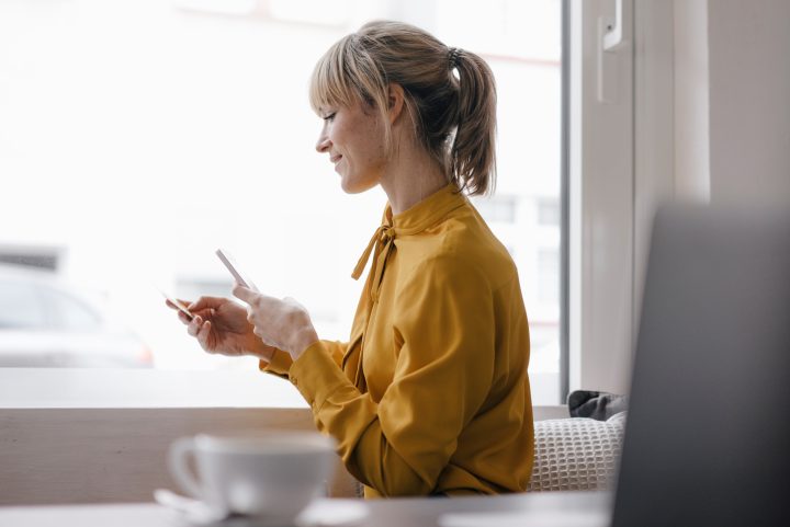 Blond businesswoman sitting at window, doing a paymant with smartphone and creditcard