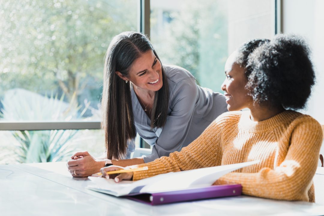 Female teacher leans on desk and smiles at female student