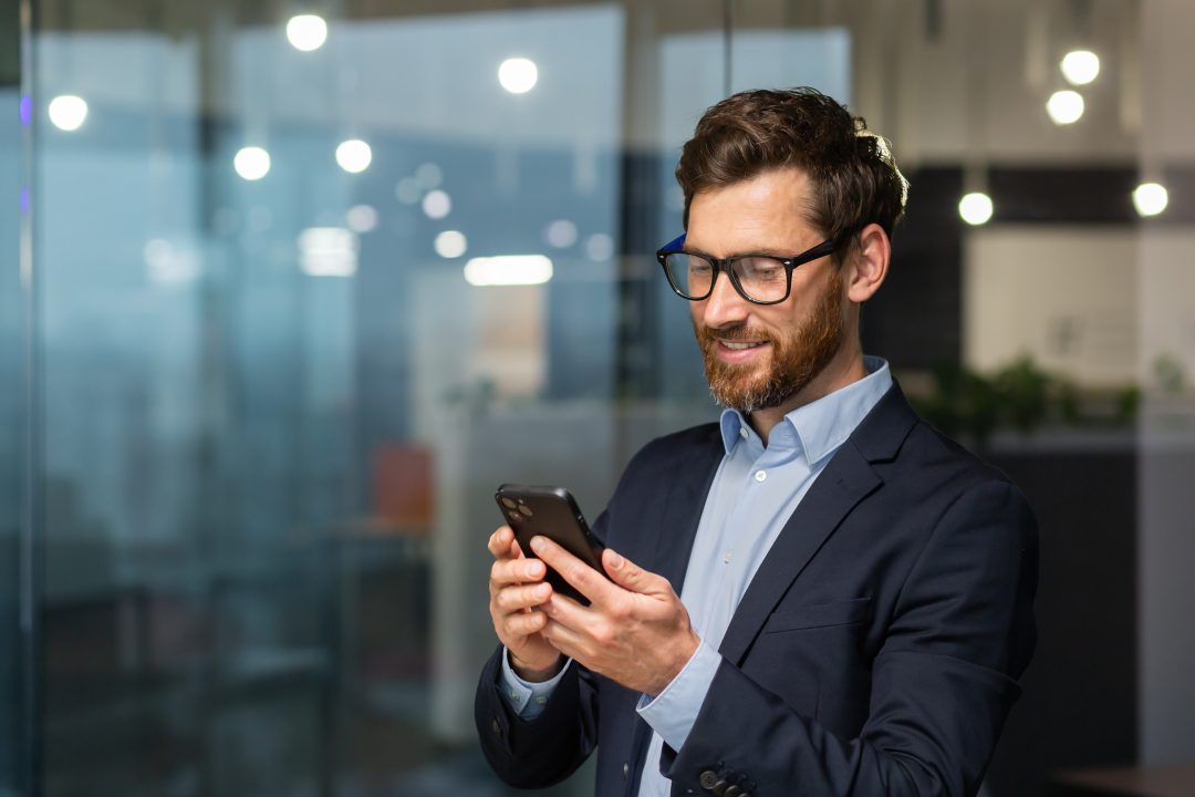 Successful financier investor works inside office at work, businessman in business suit uses telephone near window, man smiles and reads good news online from smartphone